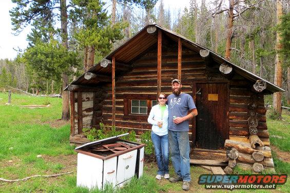 Couple standing in front of a rustic log cabin in a wooded area. An old white stove sits in the foreground