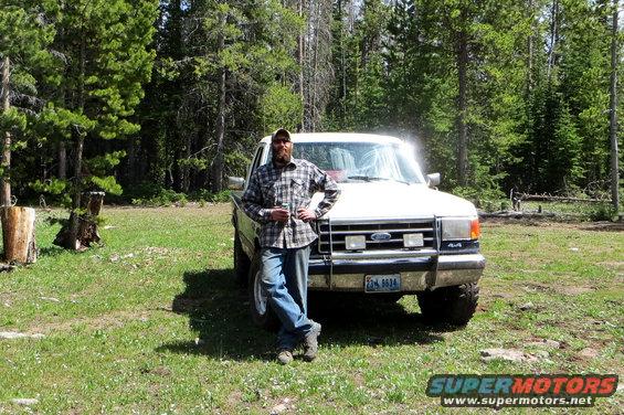 Guy leaning on a white Ford F-Series pickup, looks like late 80s model. It's parked in a grassy, wooded area