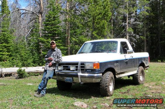 White and dark blue Ford F-Series truck with off-road tires parked on grassy terrain near trees
