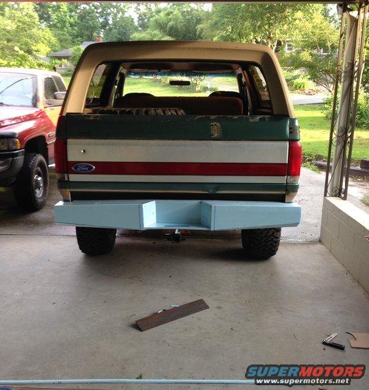 Rear view of a Ford Bronco with a light blue custom bumper. A red pickup truck is parked to the left