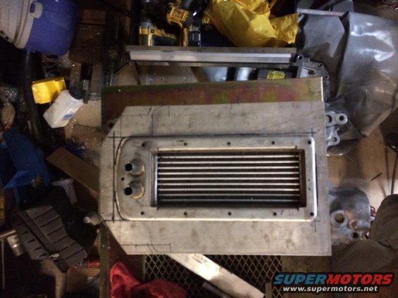 Aluminum intercooler with fins and two inlet ports, sitting on a workbench with tools and parts in the background