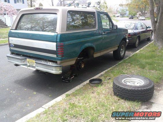 1996 Ford Bronco on a jack with a flat tire, spare tire on the ground next to it. Another car is parked behind it
