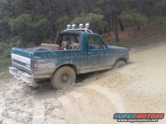 Mud-covered green Ford Bronco stuck in a dirt track, rear-left wheel submerged