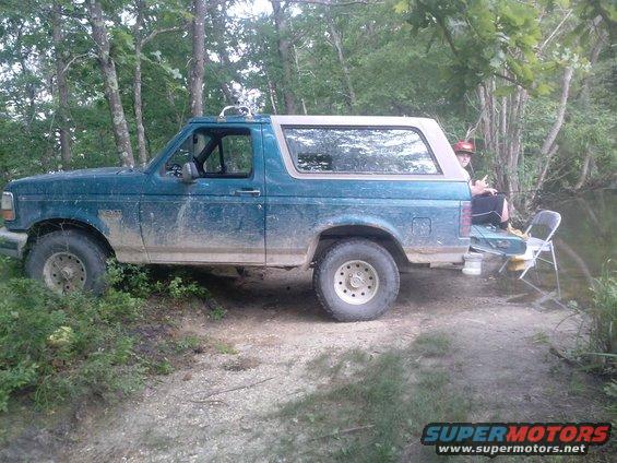 1996 Ford Bronco parked on dirt near water, person sitting on tailgate