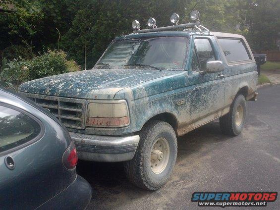 Green Ford Bronco covered in mud, parked next to another car. It has a roof rack with lights