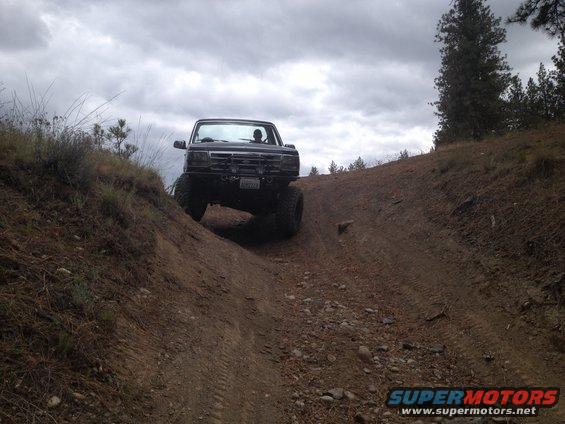 Black truck with large tires driving up a dirt hill under a cloudy sky. License plate is visible