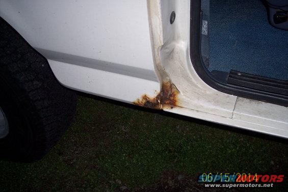 White vehicle's lower door frame showing significant rust damage near the wheel well. Interior blue carpet visible