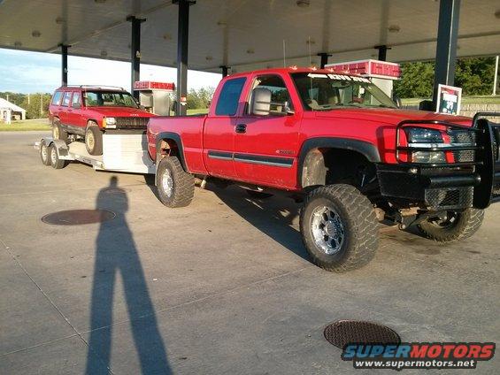 Red pickup truck towing a red Jeep Cherokee on a trailer at a gas station