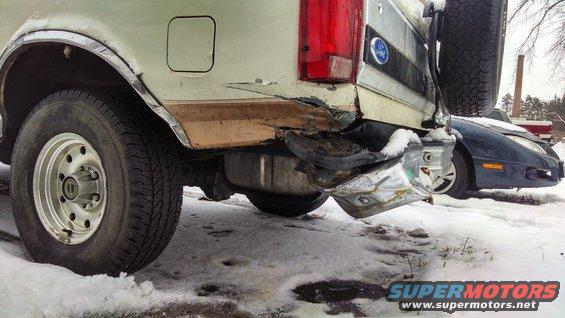 Damaged rear of a light-colored Ford Bronco with a spare tire mounted on the back. A blue sports car is visible behind it