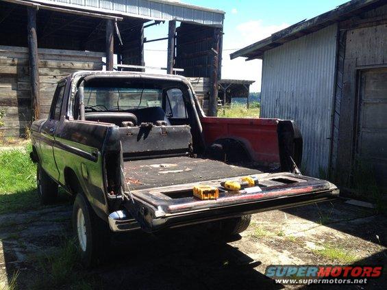 Rear view of a black Ford Bronco with its tailgate open, showing the bed and some tools. It's parked next to a weathered barn