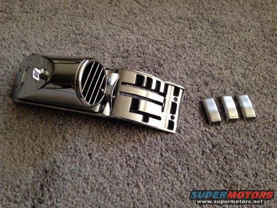 Chrome vent trim piece with horizontal slats and three small chrome emblems on a carpeted surface