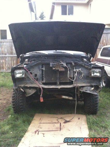 Truck front end with hood up, radiator exposed, wires hanging. Another truck is parked to the right