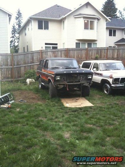 Two Ford Broncos in a yard, one with its grille removed. The Bronco in the foreground is black with orange stripes