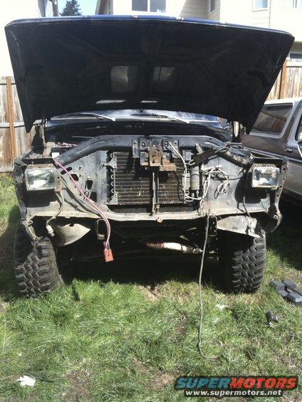 Front of a truck with hood up, grille removed, and headlights exposed. Another vehicle is partially visible to the right