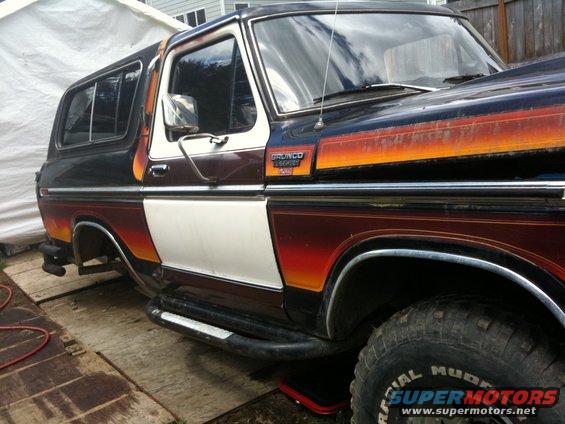 Side view of a dark blue, white, and orange striped Ford Bronco with a removable top. The front fender and door are visible