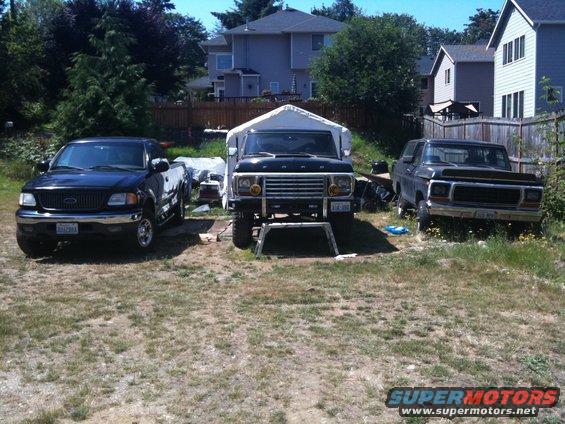 Black 1978 Ford Bronco on jack stands under a canopy, front view. Another black Ford truck is to the left, and a dark gray Ford truck is to the right