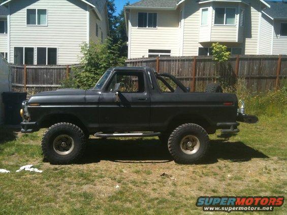 Side view of a black Ford Bronco with off-road tires and a roll bar. It's parked in a grassy yard with houses in the background