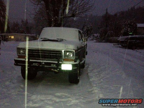 Dark green Ford Bronco covered in snow, front view. Headlights are on
