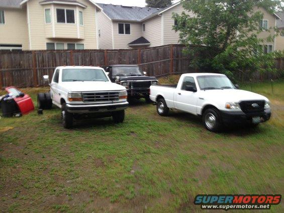White Ford pickup, black pickup, and white Ford Ranger pickup parked in a grassy yard with houses in the background