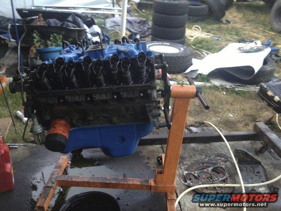 Engine on an orange stand, with blue oil pan and black valve covers. Tires and other parts are in the background
