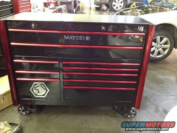 Black and red Matco tool chest with silver emblem on wheels in a garage with a car in the background
