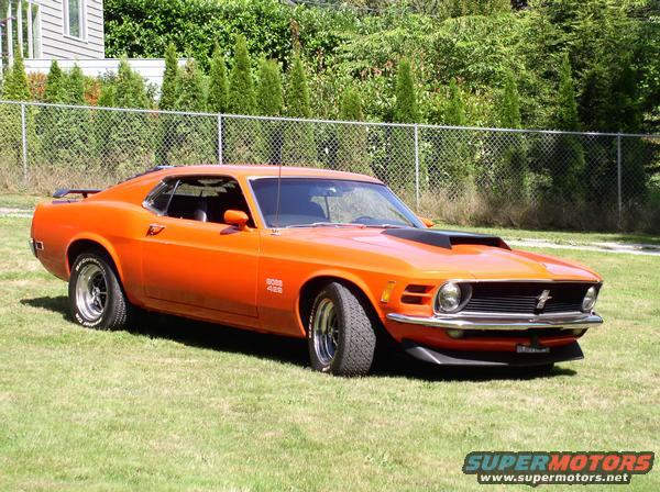Orange 1970 Ford Mustang Boss 429 parked on grass, front 3/4 view. Black hood scoop, black front splitter