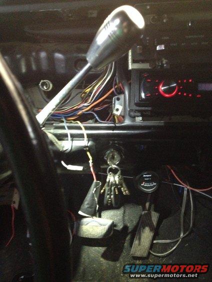 Car interior showing dashboard, steering column, gear shifter, exposed wiring, and pedals. Aftermarket radio installed