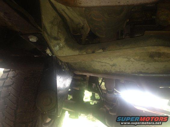 Underside of a vehicle with exposed frame, suspension components, and a tire. A watermark reads SUPERMOTORS.NET