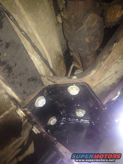 Underbody view showing a black bracket bolted to frame rails, with a driveshaft and U-joint visible