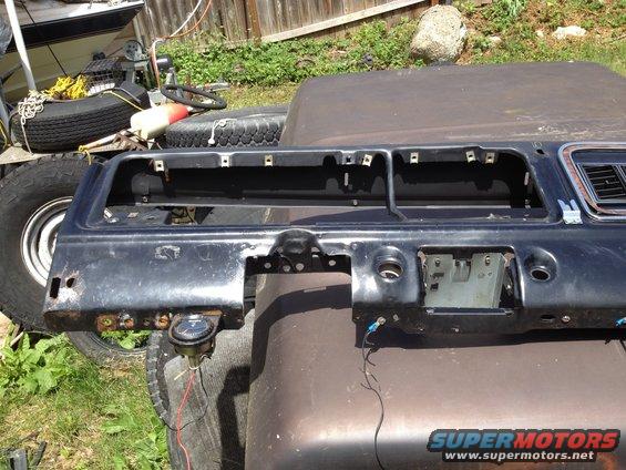 Black dashboard section with gauge and wiring, resting on a brown surface. Spare tires and boat in background