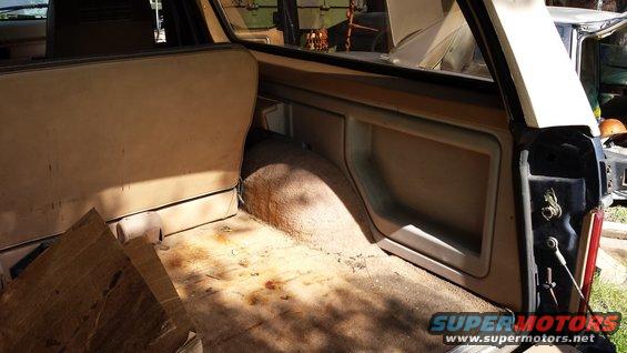 Interior cargo area of an older SUV, showing carpeted wheel wells and a dirty floor with some wood planks