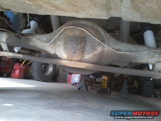 Underside view of a vehicle's rear axle, differential, shocks, and brake lines. Tires and shop equipment are visible in the background