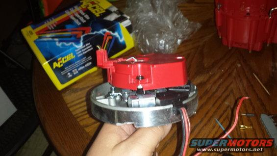 Hand holding a red automotive distributor cap and rotor assembly. Box for ACCEL spark plug wires is in the background