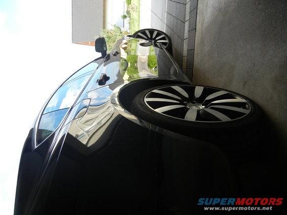 Black car, three-quarter front view, parked next to a concrete wall. Features multi-spoke alloy wheels