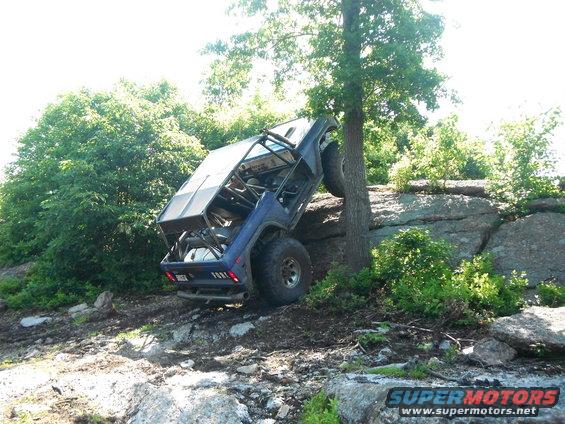 1988 Ford Bronco modified for off-roading, tilted on its side against a tree on a rocky incline