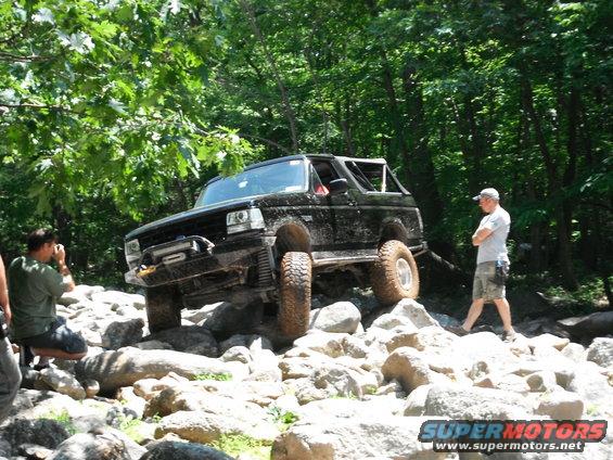 Black Ford Bronco climbing over rocks on a trail. A person is taking a photo of it