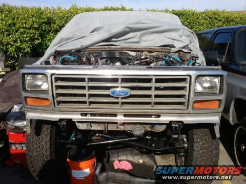 Ford truck front end, hood up, engine exposed, covered with a tarp. Another truck is parked to the right