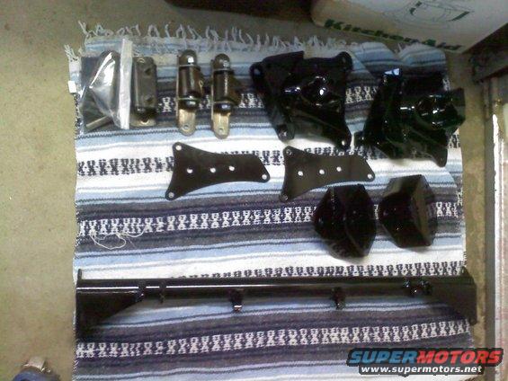 Various black metal automotive parts laid out on a striped blanket. Includes brackets, hinges, and a long bar