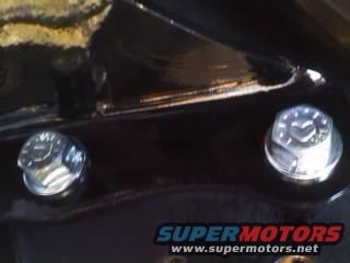 Two hex bolts with visible markings on a black painted surface. Supermotors.net logo visible