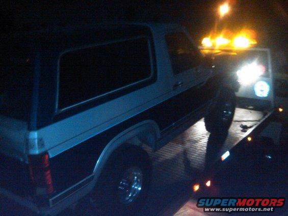 Two-tone blue and white Ford Bronco on a tow truck at night