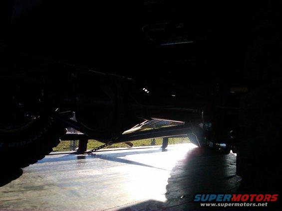 Underside view of a vehicle, showing the rear axle and suspension components. A chain is visible securing the vehicle