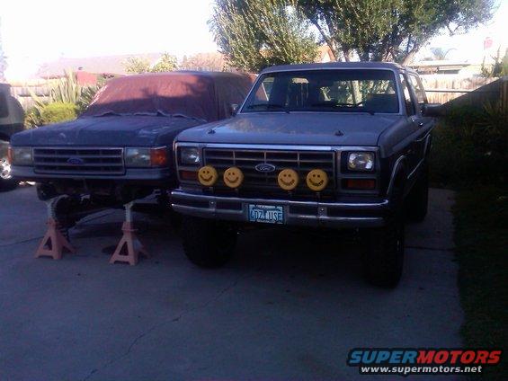 Two Ford Broncos parked outside, one in front with smiley face lights, the other behind it on jack stands