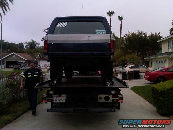 Guy in uniform walks next to a dark blue Ford Bronco on a flatbed tow truck. The Bronco has a silver stripe and a number 390 on the rear window