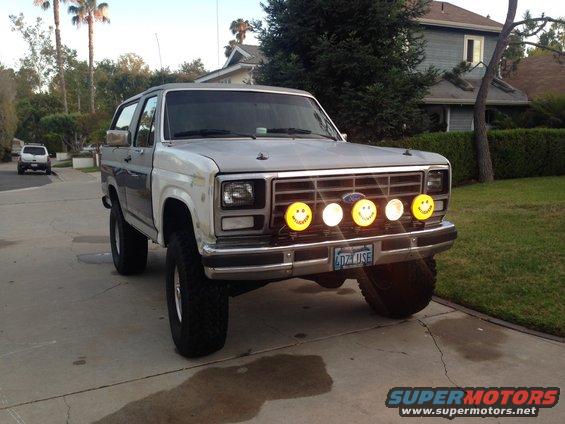 Gray and white Ford Bronco with smiley face fog lights and off-road tires parked in a driveway