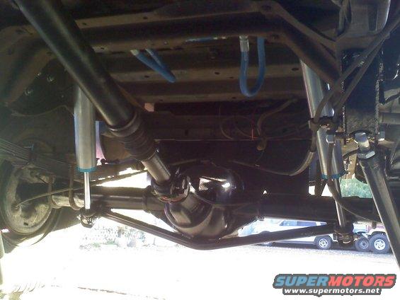 Underside view of a vehicle's rear axle, driveshaft, and suspension with blue hoses. Another vehicle is visible in the background