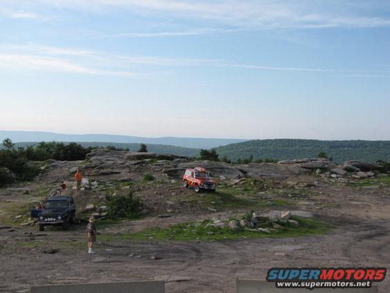 Two off-road vehicles parked on a rocky hill with people standing around. One is red, the other is dark blue