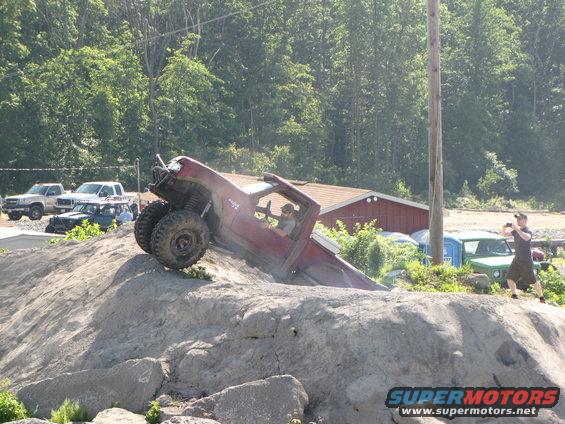 Red off-road truck tilted on a rocky incline, with large tires and a roll cage