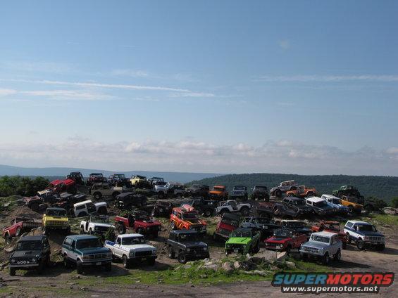 Many off-road vehicles parked on a rocky hillside under a blue sky
