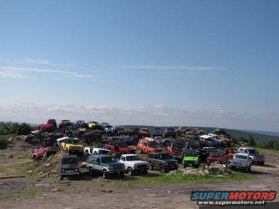 Many SUVs and trucks parked on a grassy hill under a blue sky