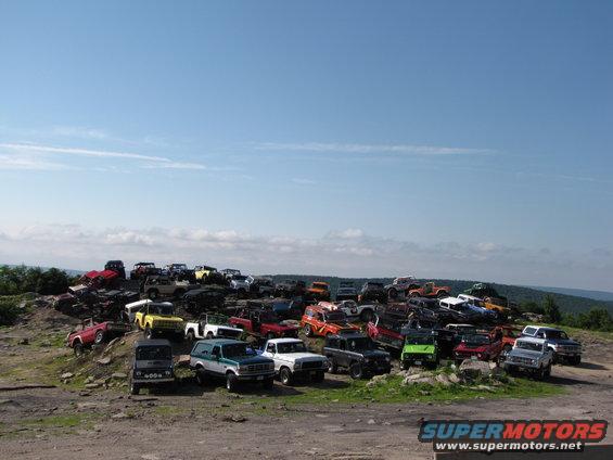 Many off-road vehicles parked on a rocky hill under a blue sky